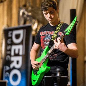 Person playing a green electric guitar with a Derosa Music sign in the background
